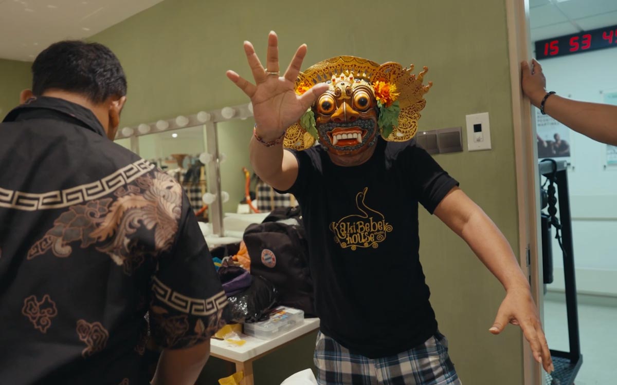 A male performer wearing a traditional Balinese mask is gesturing at the camera in the dressing room, backstage at Esplanade. 